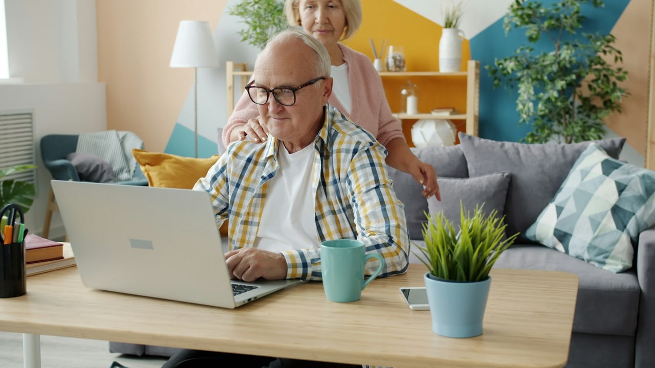 An older man using a laptop at home while his wife stands beside him